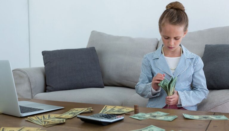 Young girl counting cash at a table with a calculator, laptop, coins, and bills.