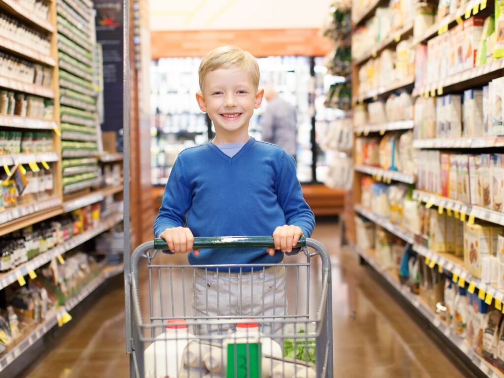 Smiling boy in a blue sweater pushes a shopping cart with groceries down a brightly lit grocery aisle, surrounded by shelves full of packaged foods.