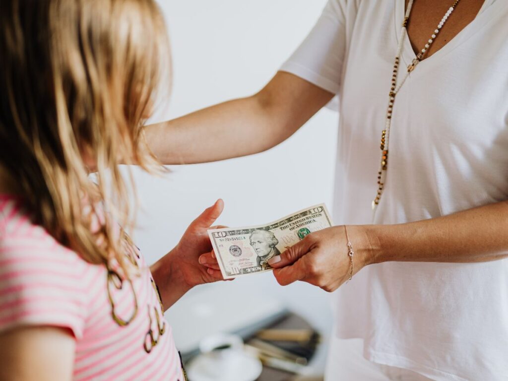 A woman in a white shirt hands a ten-dollar bill to a child wearing a pink striped shirt.