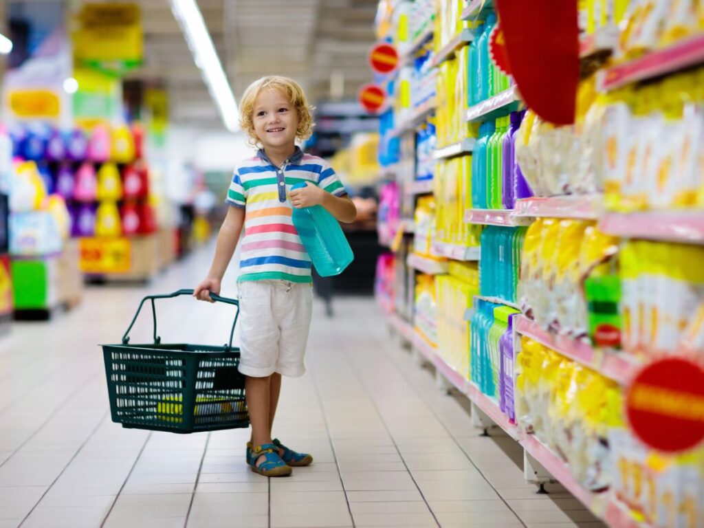 Young child in a striped shirt and shorts smiles while holding a basket and bag in a colorful supermarket aisle