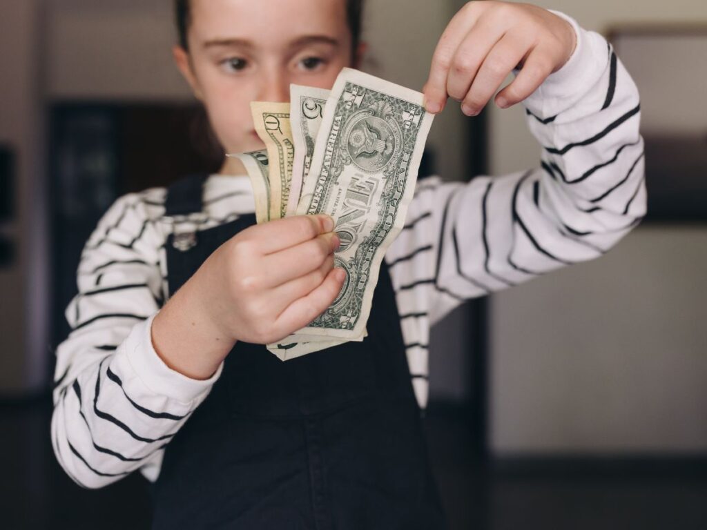 A child in a striped shirt holds several U.S. dollar bills, including a one and a five, with a focused expression.