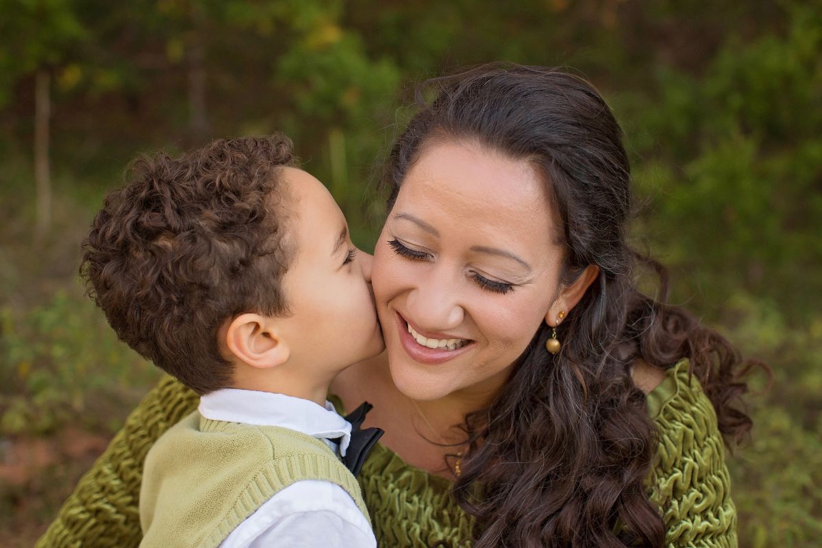 Mom smiling as her young son kisses her on the cheek outdoors