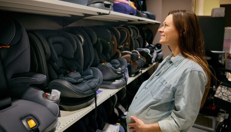 Pregnant woman shopping for a child car seat in a store with rows of car seats on display.