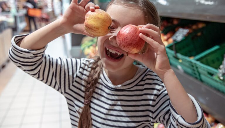 Young girl playfully holding two apples over her eyes while shopping in a grocery store.