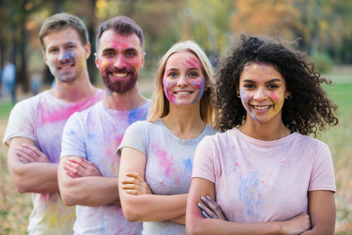 Four friends standing outdoors with pink and blue powder on their clothes and faces, celebrating together.