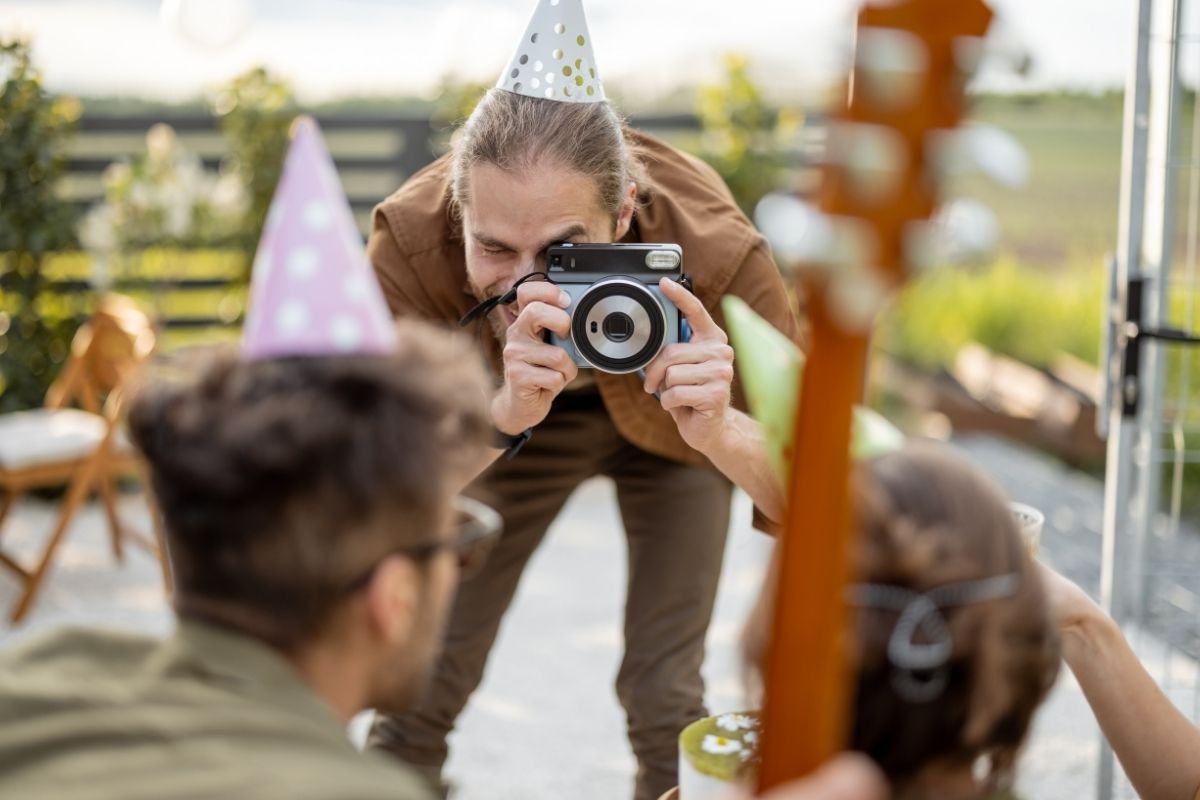 Man wearing a party hat taking photos of guests at an outdoor celebration.