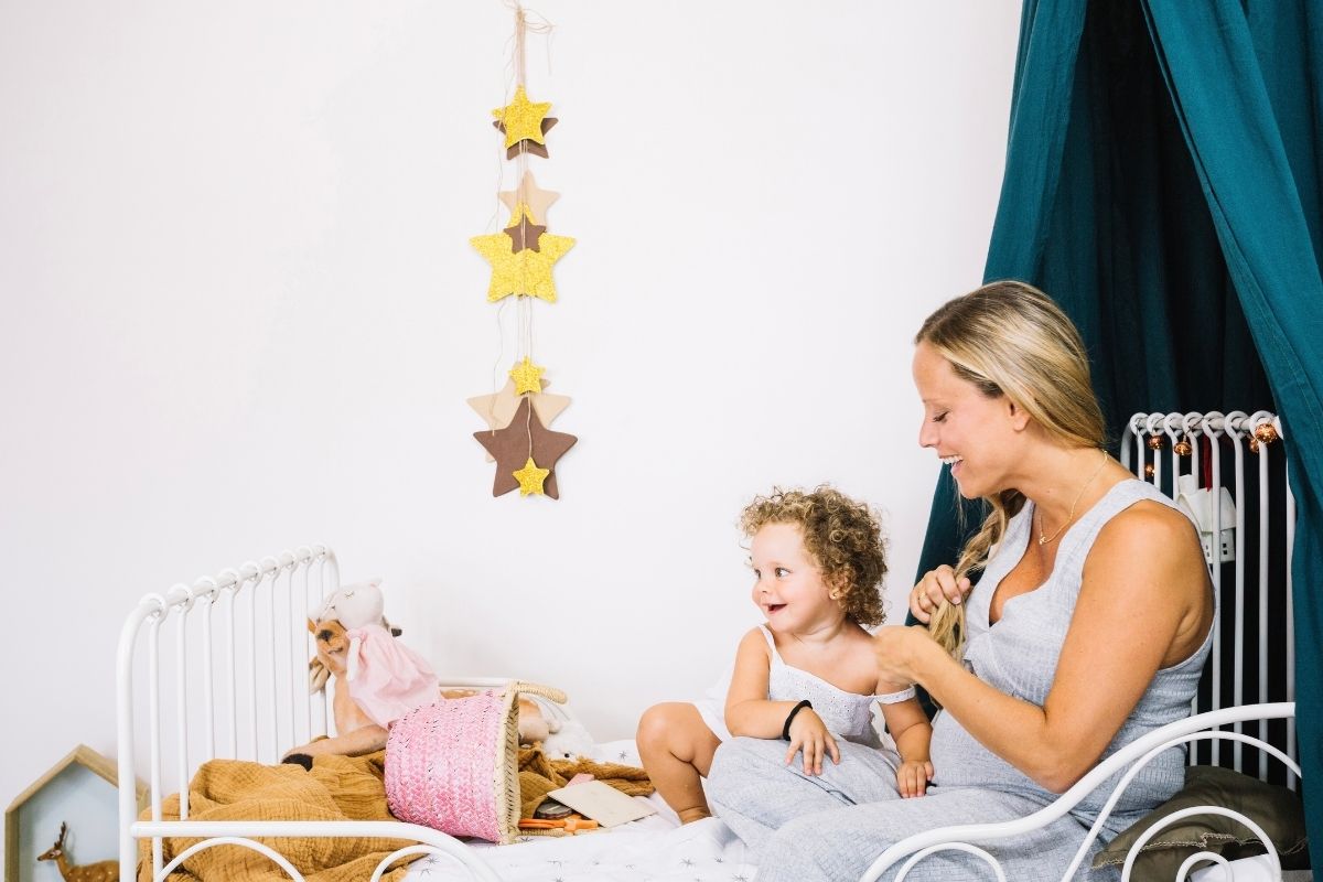 A mother sitting beside her smiling toddler on a small white bed in a cozy children's bedroom decorated with hanging stars, toys, and a dark teal canopy.